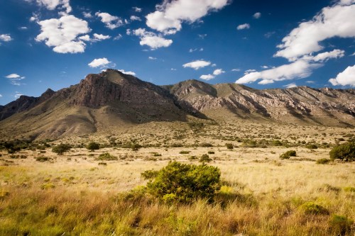 A vast landscape featuring rolling hills, rocky mountains, and a blue sky with scattered clouds.