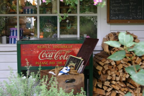 A vintage Coca-Cola cooler beside a stack of firewood, with a sign inviting customers to serve themselves.