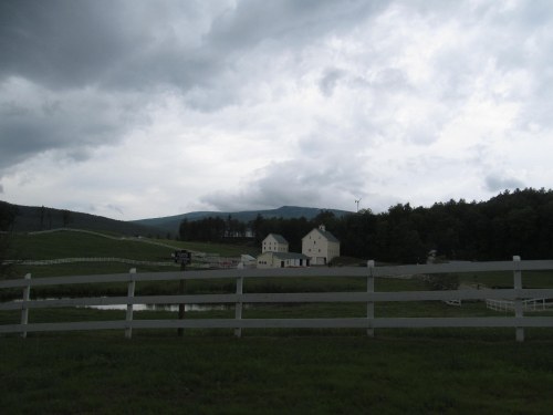 A cloudy sky looms over a rural landscape with white houses and a fence, surrounded by green fields and distant hills.