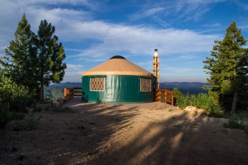 A green yurt on a wooden deck surrounded by trees, with mountains in the background under a blue sky.