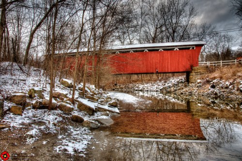 A red covered bridge over a stream, surrounded by bare trees and rocky banks, with a hint of snow on the ground.