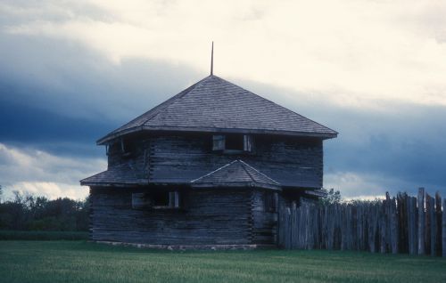 A historic wooden structure with a peaked roof, surrounded by a wooden fence, set against a cloudy sky.