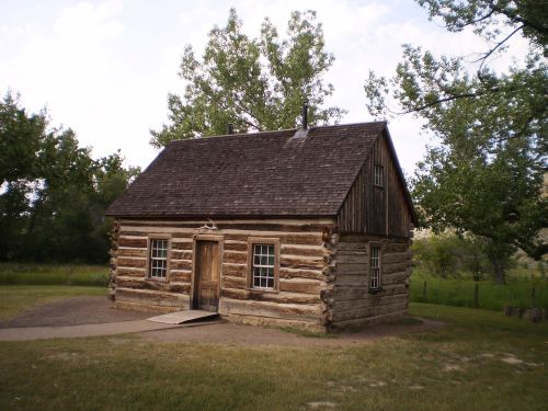 A rustic log cabin with a sloped roof, surrounded by trees and grass, set against a clear sky.