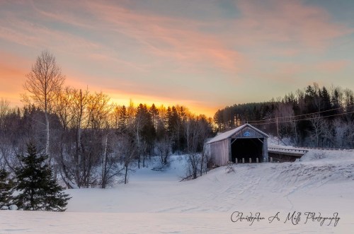 A serene winter landscape featuring a covered bridge at sunset, surrounded by snow and trees.