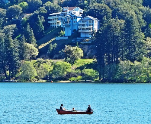 A small boat with three people floats on a blue lake, surrounded by lush greenery and hillside homes.