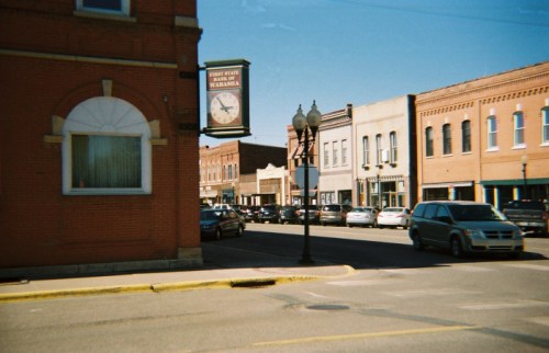A street view of a small town with brick buildings, parked cars, and a clock sign on the corner. Clear blue sky above.