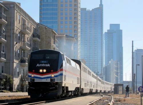 An Amtrak train travels through a cityscape with tall buildings in the background on a clear day.