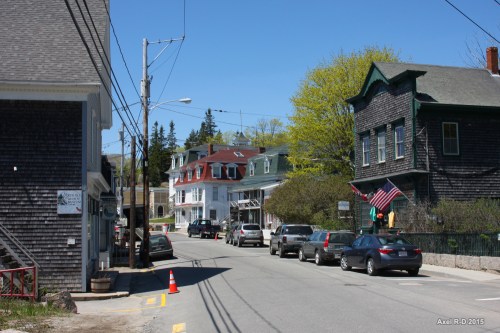 A quiet street scene featuring historic houses, parked cars, and trees under a clear blue sky.