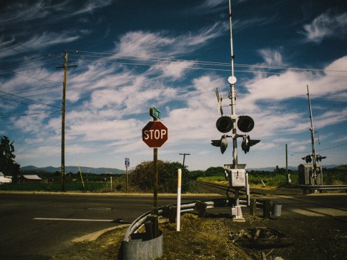 A stop sign at a railroad crossing with signals, surrounded by fields and a blue sky with clouds.