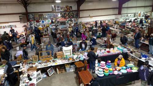 A bustling indoor market with numerous vendors and shoppers browsing various antiques and collectibles.