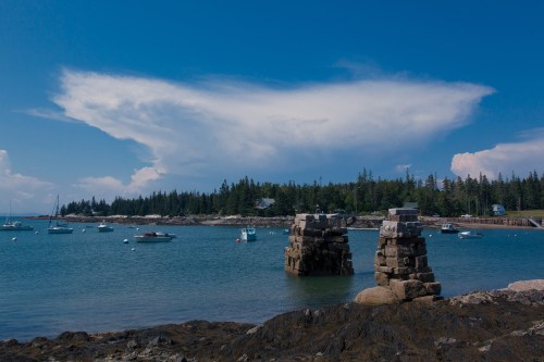 A serene coastal scene with boats in the water, rocky shore, and a dramatic cloud formation in a blue sky.