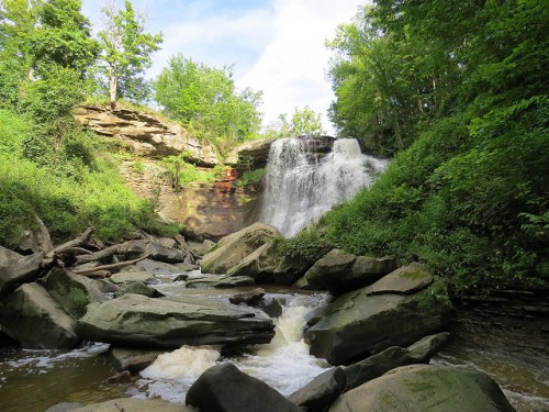 A serene waterfall cascades over rocky cliffs, surrounded by lush greenery and a gentle stream below.
