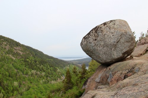 A large boulder perched on a rocky ledge overlooking a lush green valley and distant water.