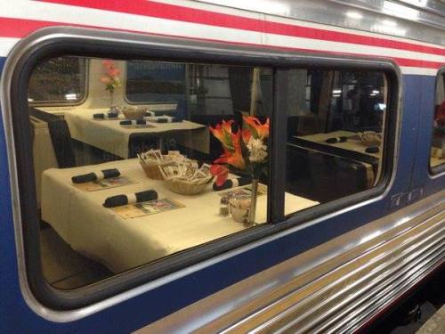 A dining car with neatly set tables, flowers, and utensils visible through a window.