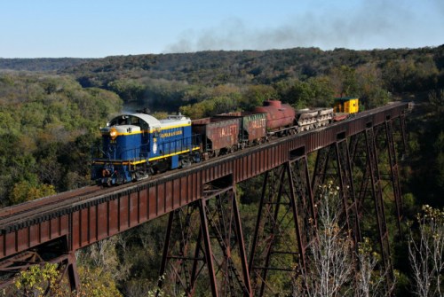 A blue and yellow train crosses a rusted bridge over a lush, green valley with smoke rising in the background.