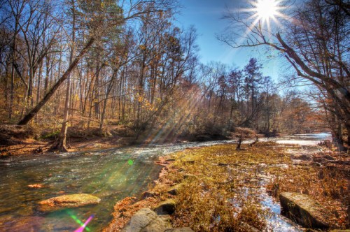 A serene river flows through a forest, with sunlight shining and autumn foliage reflecting on the water's surface.