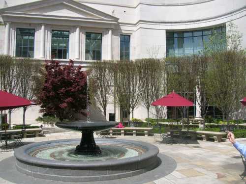 A tranquil courtyard featuring a fountain, surrounded by trees and red umbrellas over seating areas.