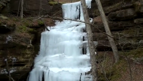 A frozen waterfall cascading down rocky cliffs, surrounded by bare trees and a forested landscape.