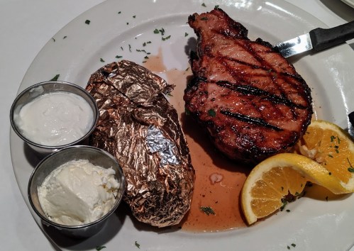 A grilled pork chop with a baked potato, sour cream, and orange slices on a white plate.