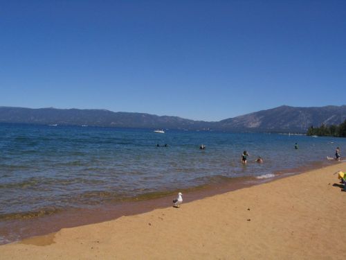 A sunny beach scene with people swimming and relaxing by the water, surrounded by mountains and clear blue skies.