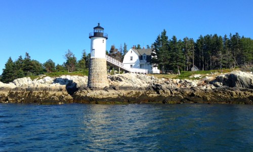 A lighthouse stands on rocky shore, surrounded by trees and a clear blue sky, with calm water in the foreground.