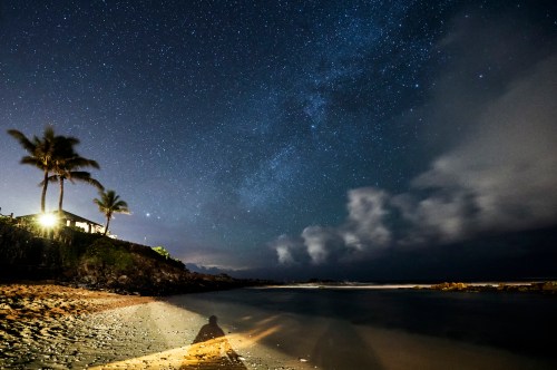 A serene beach at night, with a starry sky and clouds, illuminated by a distant light and a shadow on the sand.