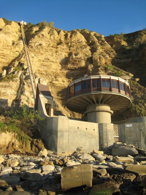 A unique circular building at the base of a cliff, with a steep lift leading up to the top.