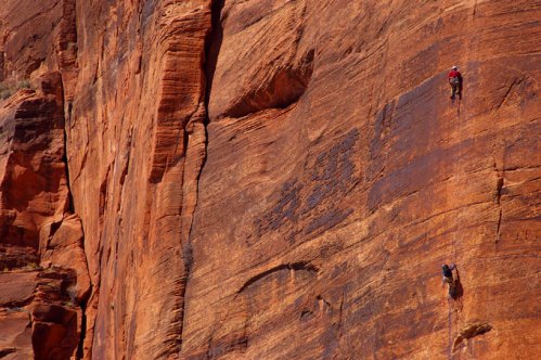 Two climbers ascend a steep, reddish rock face in a rugged outdoor setting.