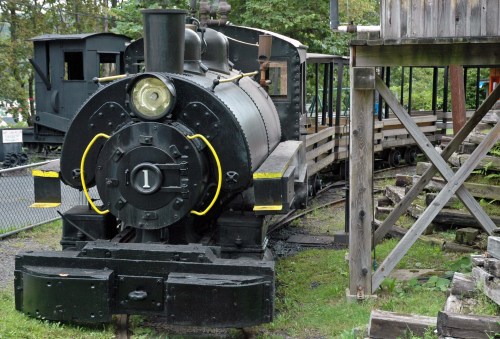 A vintage black steam locomotive with a yellow trim, parked beside a wooden platform in a green outdoor setting.
