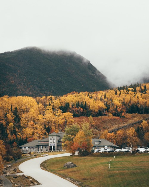 A scenic view of a house surrounded by autumn foliage and mountains, with misty clouds overhead.