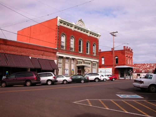 Historic buildings line a street, featuring red brick facades and parked cars under a cloudy sky.