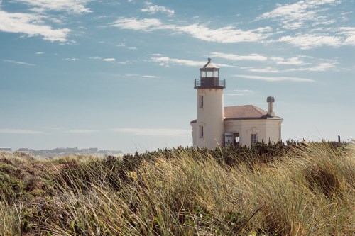 A lighthouse stands on a grassy dune under a blue sky with scattered clouds.