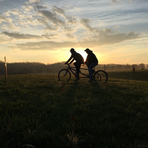 Two cyclists ride a tandem bike against a sunset backdrop, with rolling hills and a cloudy sky in the background.