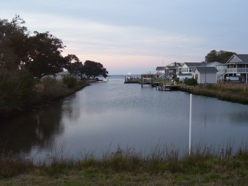 A calm waterway lined with houses, reflecting a soft sunset sky, surrounded by greenery.