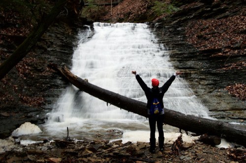 A person with a red hat stands in front of a waterfall, raising their arms in celebration amidst a forested area.