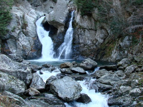 A serene waterfall cascades over rocky cliffs into a clear pool, surrounded by lush greenery and boulders.