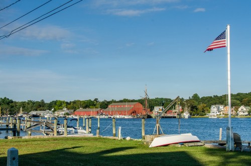 A peaceful waterfront scene with a flag, boats, and a red building in the background under a clear blue sky.