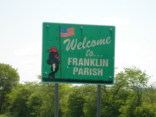 Welcome sign for Franklin Parish featuring a cartoon character and an American flag. Green background with trees in the distance.