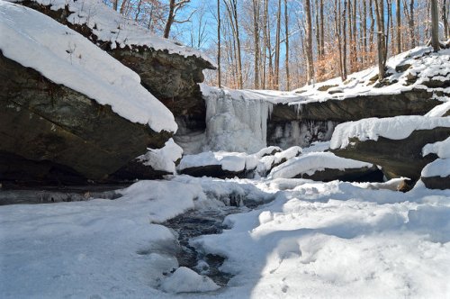 A snowy landscape featuring a frozen waterfall surrounded by rocky cliffs and bare trees under a clear blue sky.