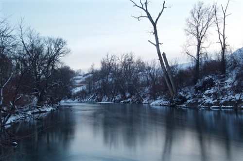 A serene winter landscape featuring a calm river surrounded by bare trees and snow-covered banks.