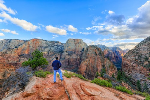 A person stands on a rocky ledge, overlooking a vast canyon with colorful cliffs and a blue sky filled with clouds.
