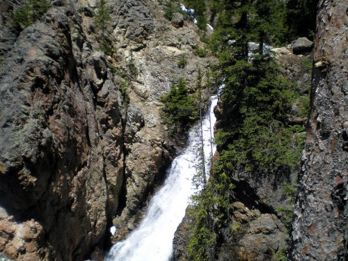 A cascading waterfall flows through rocky terrain, surrounded by lush green trees and rugged cliffs.