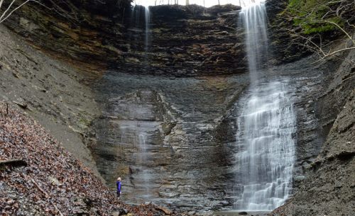 A person stands near a large waterfall cascading down a rocky cliff, surrounded by trees and a forested area.