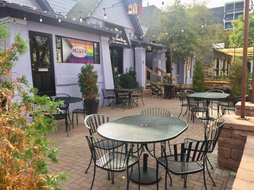 Outdoor seating area with black metal tables and chairs, surrounded by greenery and colorful banners on a purple building.