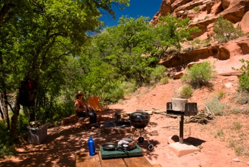 A campsite with a grill, picnic table, and a person sitting under trees near red rock formations.