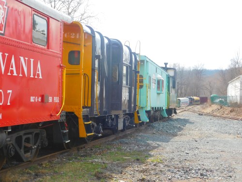 A row of colorful train cabooses parked on a gravel track, surrounded by trees and a distant train.