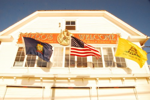 Three flags, including the U.S. flag, wave in front of a building with a sign reading "Hero's Welcome."