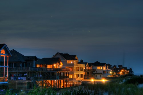 Coastal homes illuminated at night, with a sandy beach and dark sky in the background.