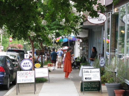 A vibrant street scene with people walking, art displays, and shops under leafy trees during an art walk event.