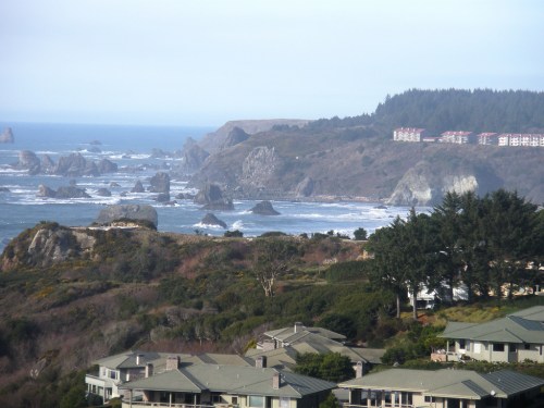 Coastal view with rocky shoreline, ocean waves, and distant hills, featuring nearby houses and greenery.